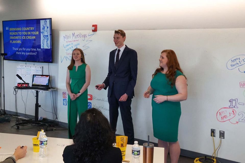 Two women wearing green on either side of one man wearing a suit presenting at a compeittion